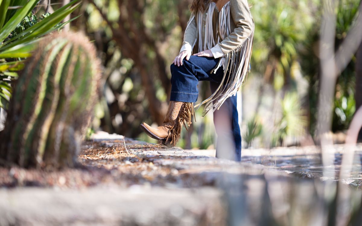 Woman in a fringe jacket and jeans posing outdoors near a cactus on a bright sunny day