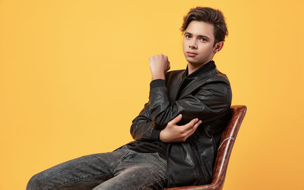 A man in a leather coat and jeans sitting with arms crossed, showing the hem line