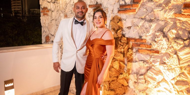 Stylish couple in formal evening attire posing together against an illuminated stone wall