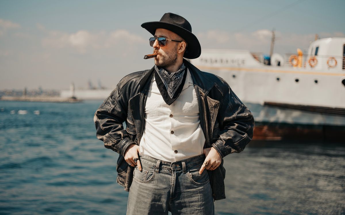 A confident man in a leather jacket standing near a boat in the sun