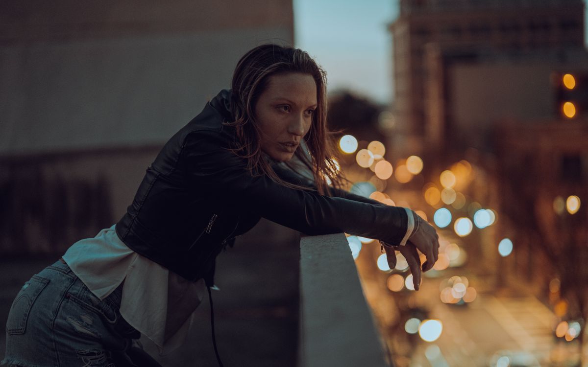 A woman in a leather jacket leaning on a rooftop ledge at twilight over a city
